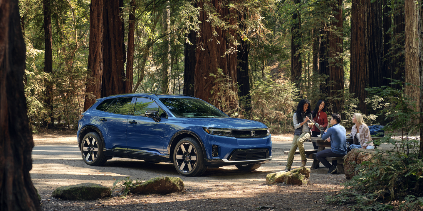 People sitting around table in the middle of woods next to 2025 Prologue vehicle.